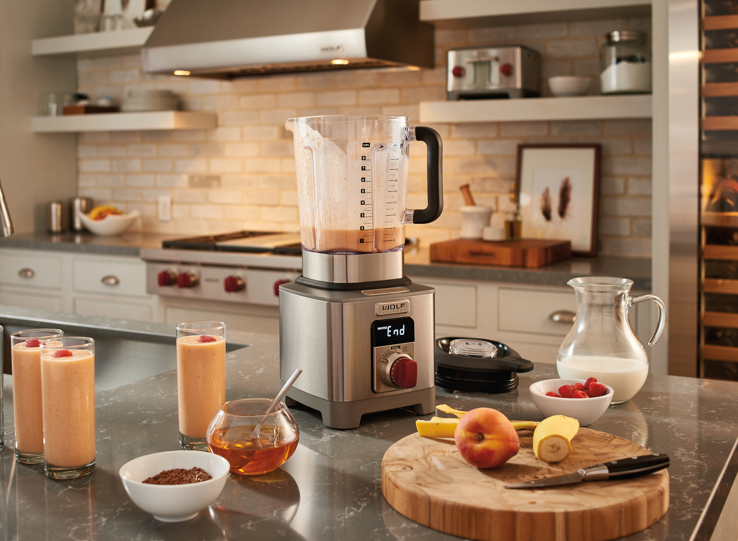 A kitchen counter with Wolf Gourmet blender and smoothies, honey and fresh fruit, and a Wolf range visible in the background.